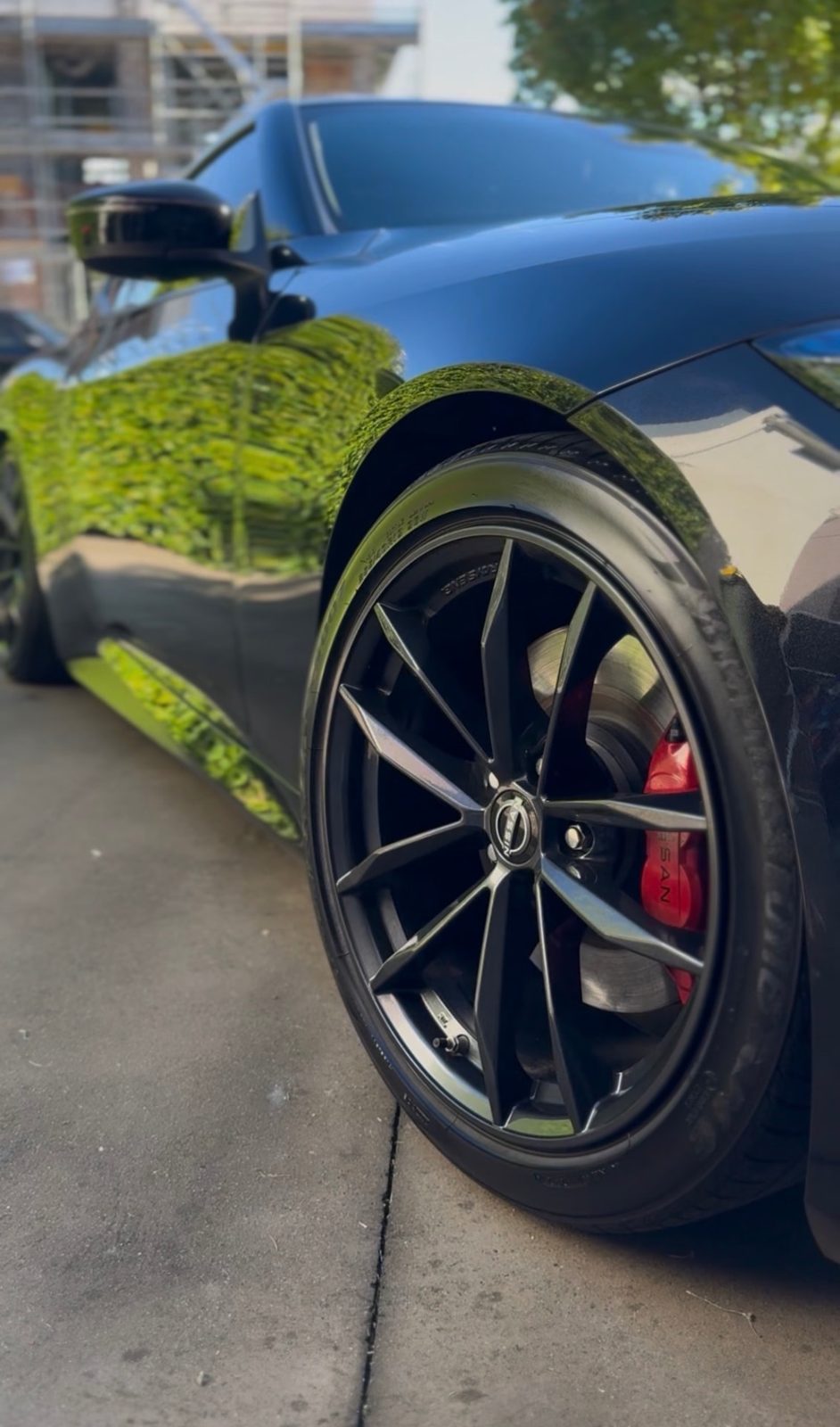 Glossy black Nissan Z side panel reflecting a green hedge, with a black wheel and red caliper